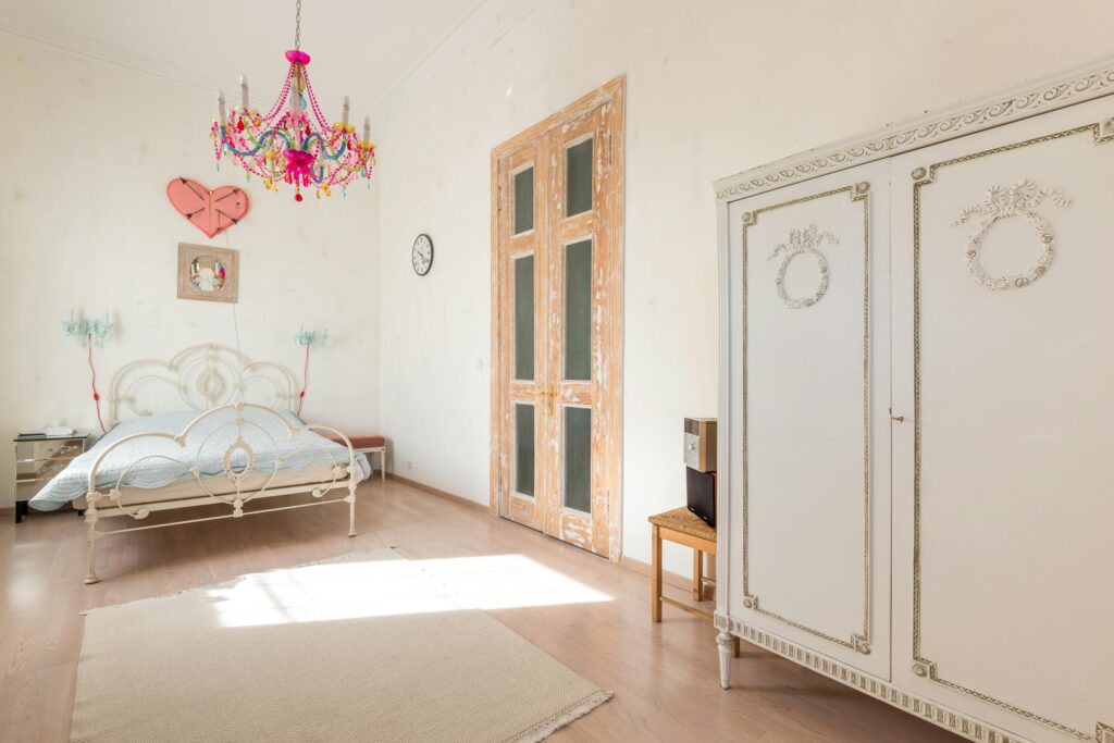 Bright vintage bedroom with ornate bed, chandelier, and wardrobe in soft light.