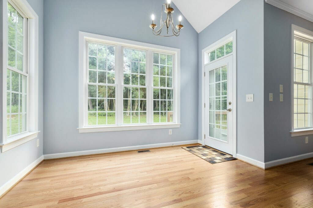 Sunlit empty room with wooden floor, chandelier, and French windows leading outside.